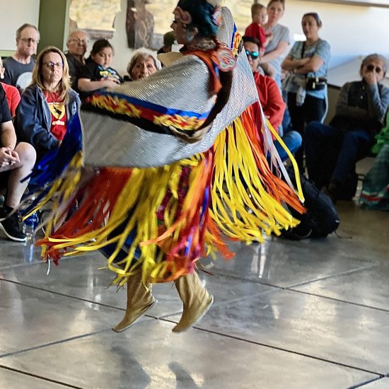 A Nuwuvi dancer with red organce and yellow ribbons and a colorfully beaded shawl dances animatedly in the hall of red rock canyon visitor center